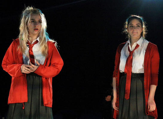 Two girls are on stage dressed in school uniform speaking to each other