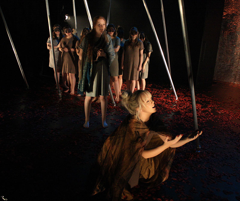 An on stage production in which a group of people are standing on stage. The girl at the front is kneeling with her arms spread widely.