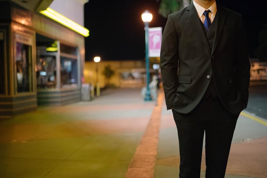 A man in a suit stands at a train station. His face is outside the frame of the photograph