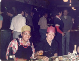 Two women in 1980s clothing and paper party hats sit in front of party food
