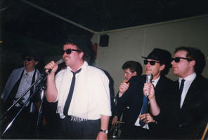 Five men wearing sunglasses and suits are singing into microphones in the 1980s