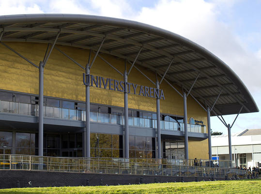 An outside shot of the University arena. It is a gold building with a sloped roof.