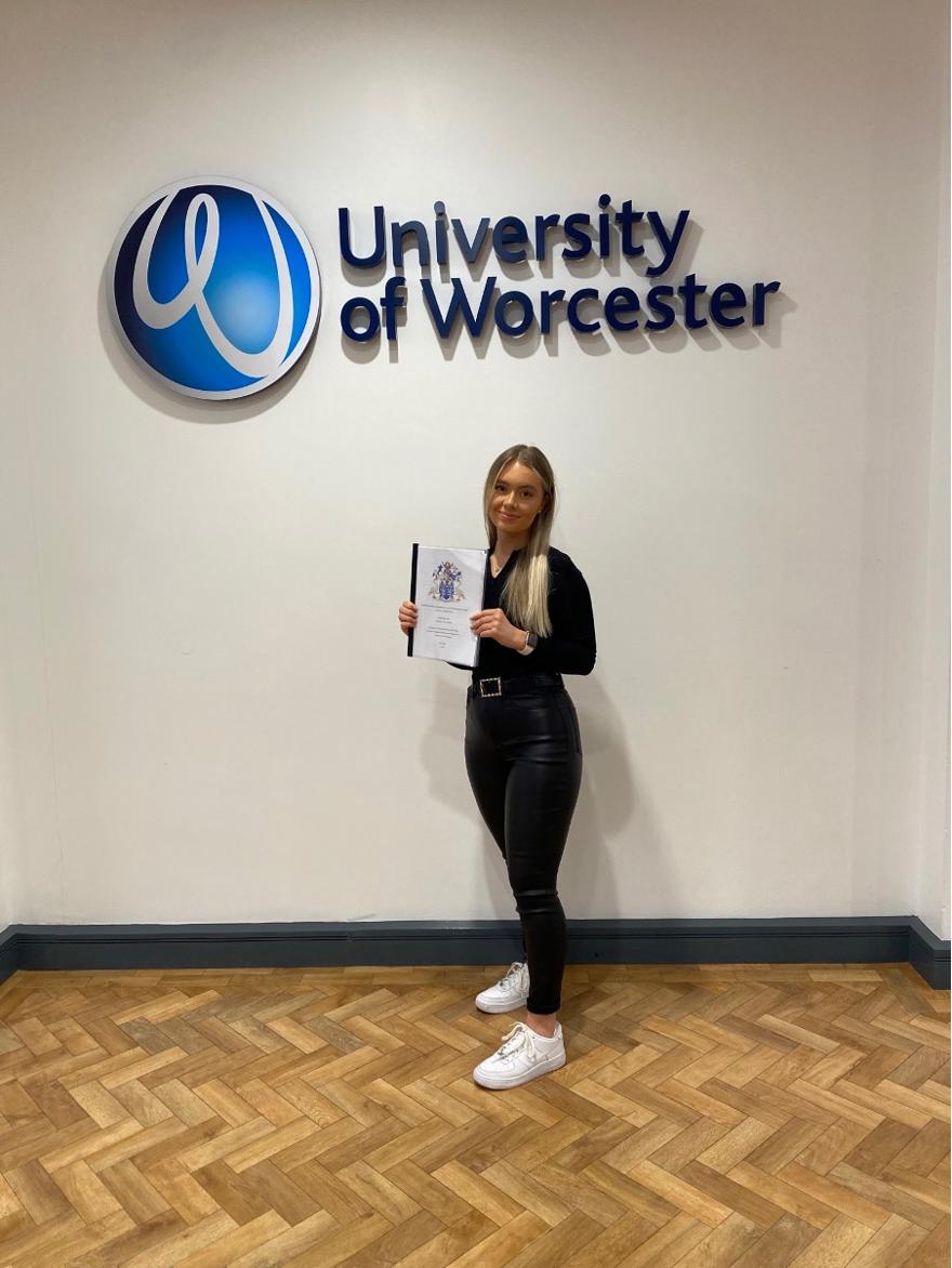 Criminology student stood in front of the University of Worcester logo holding her dissertation project