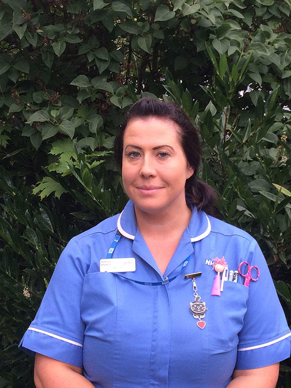 Kathryn Watson is wearing a nurse's uniform and standing against a green foliage background