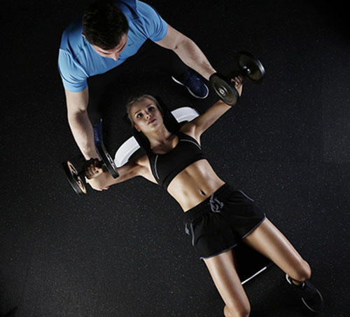 A woman is lying on a weights bench lifting two hand weights. She has a trainer spotting her.