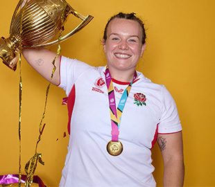 Lark wearing her England cricked team shirt, holding up her trophy and wearing a medal. 