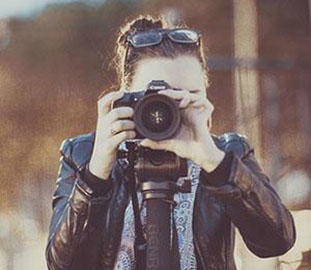 A woman with tied up brown hair and glasses on the top of her head is taking a photograph using a DSLR camera