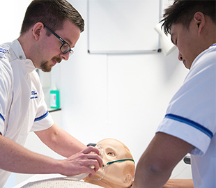 Two male student nurses supporting a dummy patient in a simulation ward